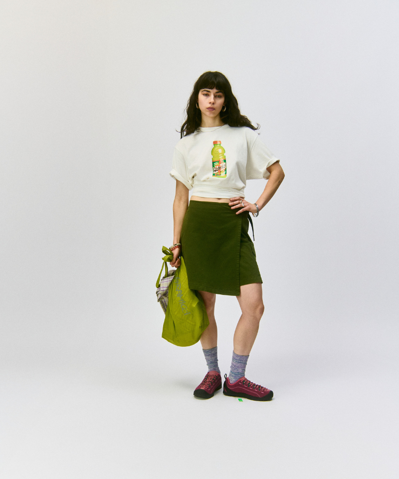 Model wearing a white shirt and a green skirt holding an olive Gramicci Ultra-Light Shopper on a light-tone background.