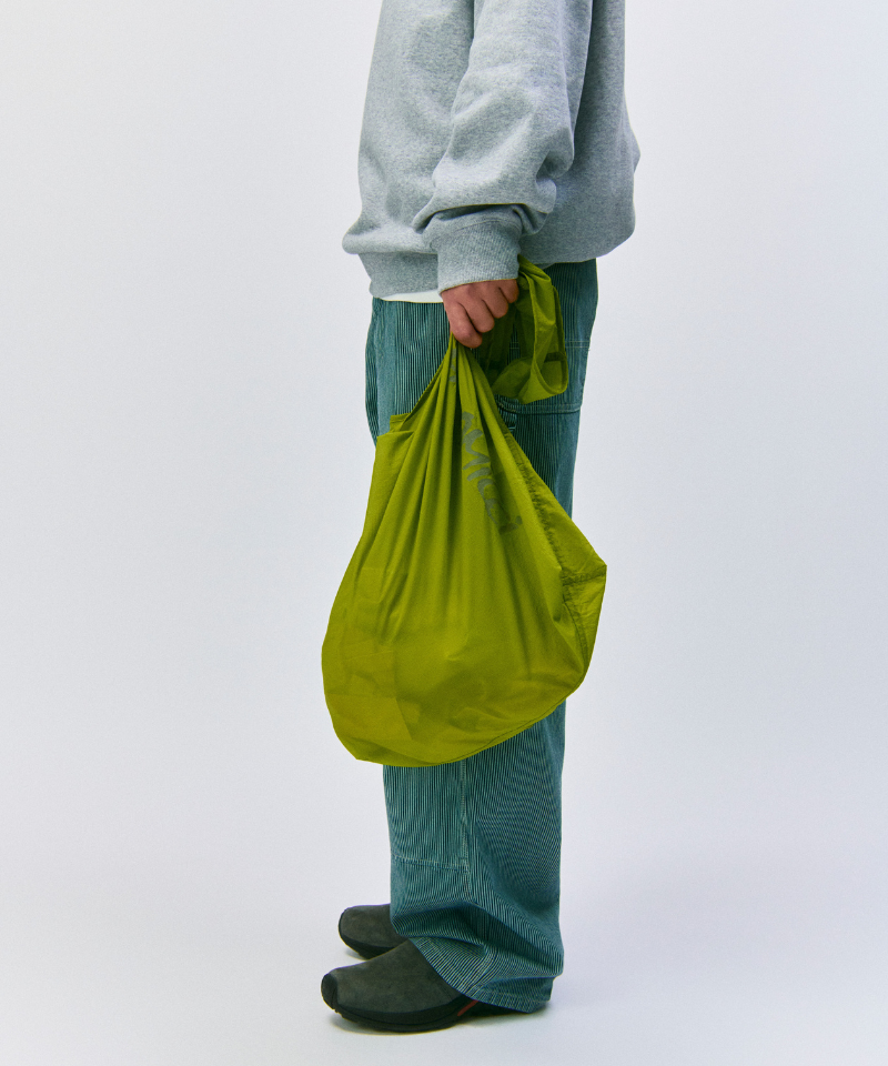 Model wearing a grey sweatshirt and green pants holding an olive Gramicci Ultra-Light Shopper on a light-tone background.