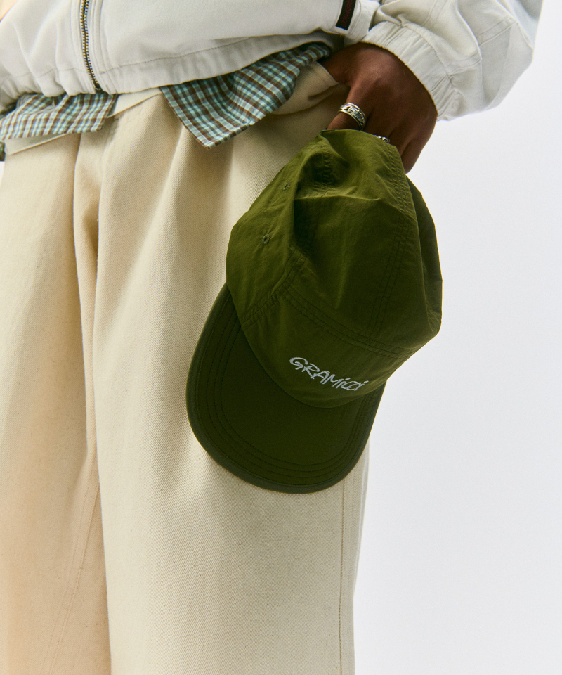 Model holding an olive Nylon Gramicci Cap on a light-tone background.