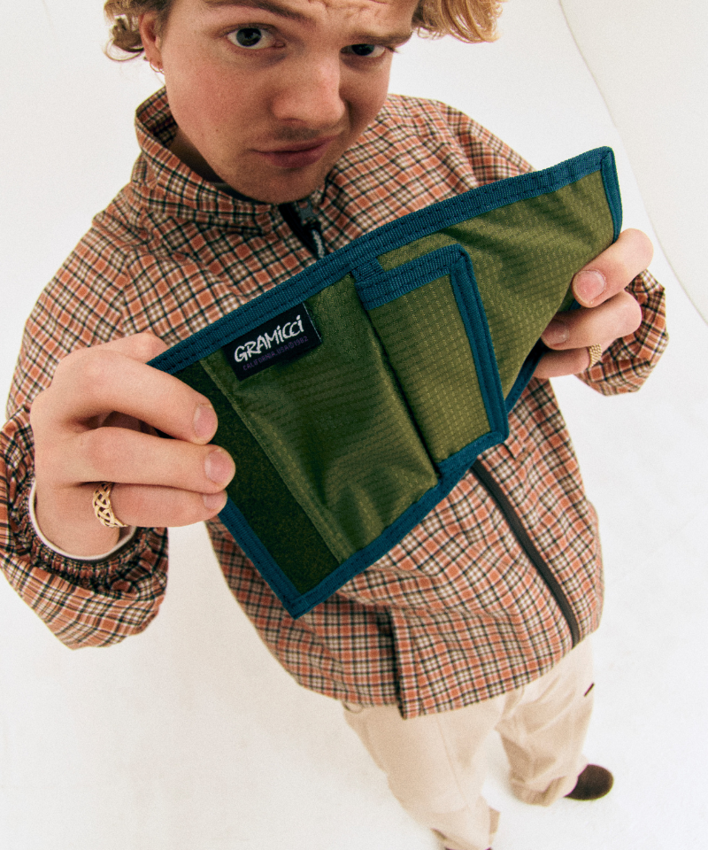 Model wearing a plaid shirt holding an olive Gramicci Velcro Wallet on a light-tone background.