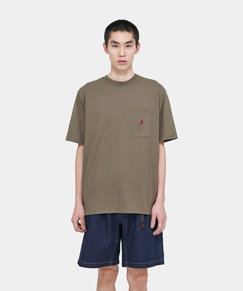 Model wearing a coyote brown Gramicci One Point Tee with a small running man graphic and blue shorts on a light-tone background.