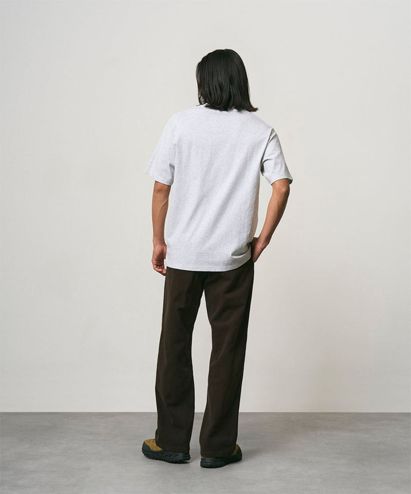 Back view of a model wearing a ash heather Gramicci One Point Tee and brown pants on a light-tone background.