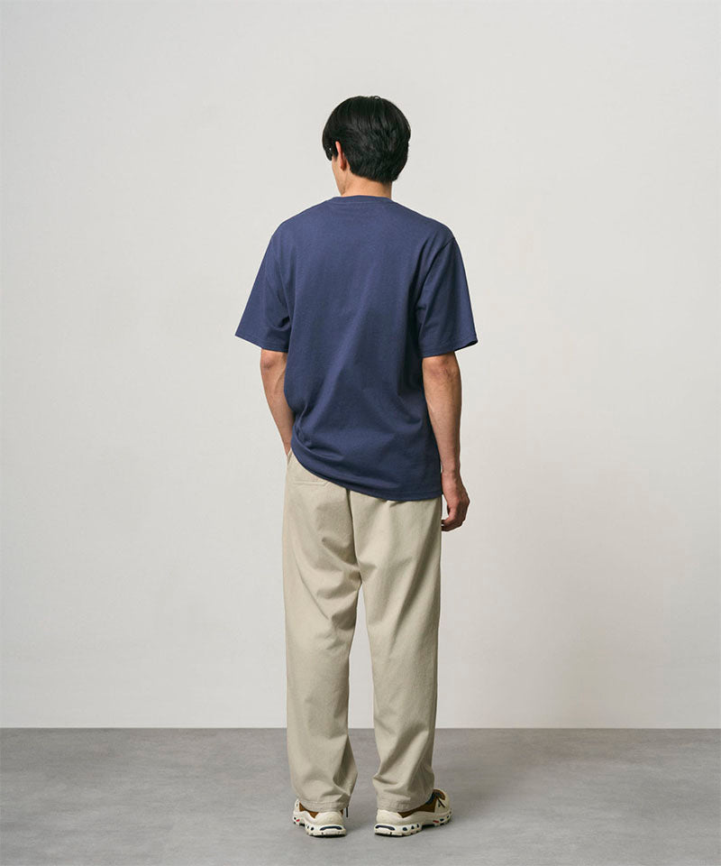 Back view of a model wearing a navy Gramicci One Point Tee and beige pants on a light-tone background.