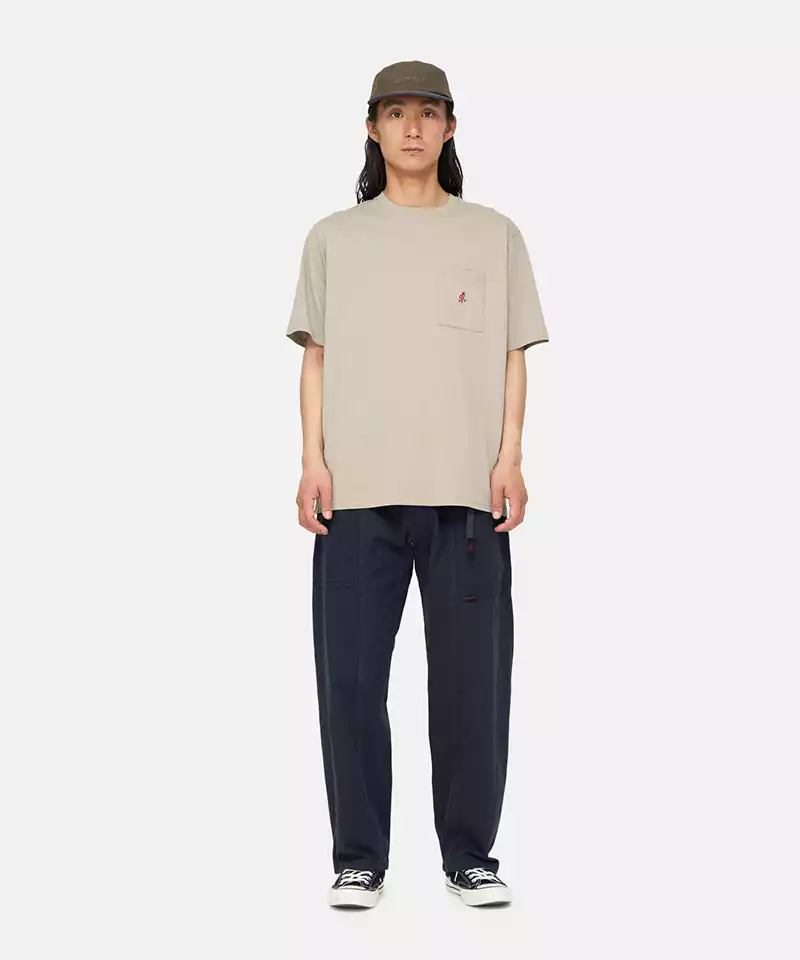 Model wearing a smoky sand colored Gramicci One Point Tee with a small running man graphic and blue pants on a light-tone background.