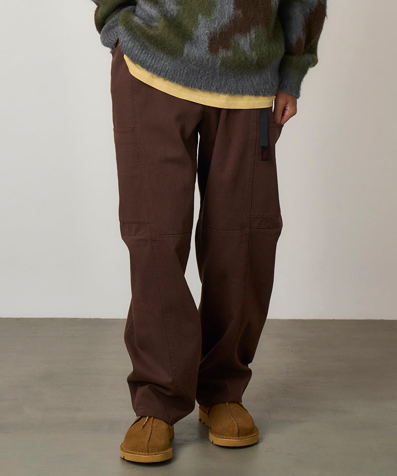 Model wearing tobacco colored Gramicci Voyager Pants and a red jacket on a light-tone background.