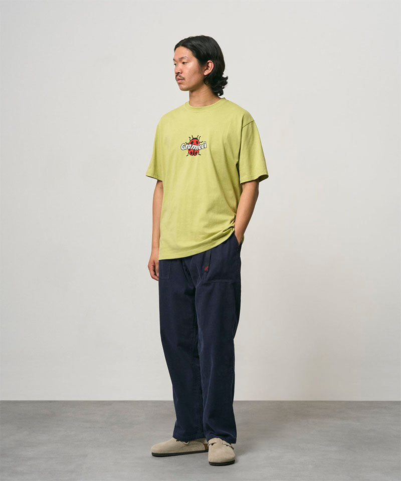 Model wearing a sweet pea colored Gramicci Ladybug Tee with colorful graphic and blue pants on a light-tone background.
