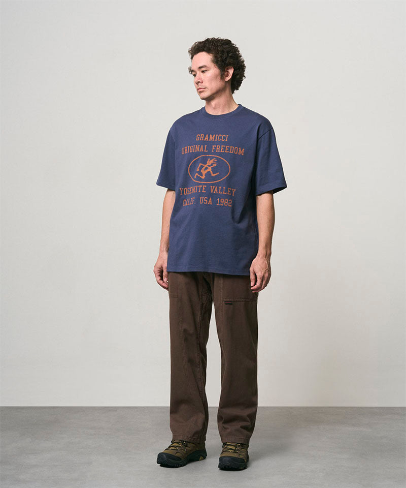 Model wearing a navy Gramicci Yosemite Valley Tee with a graphic and brown pants on a light-tone background.