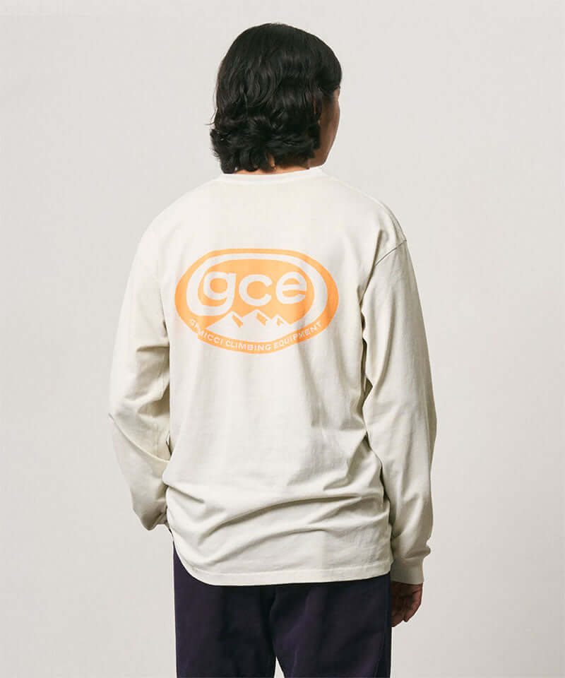 Back view of a model wearing a natural pigment colored Gramicci GCE Tee Pigment Dyed with a graphic and black pants on a light-tone background.