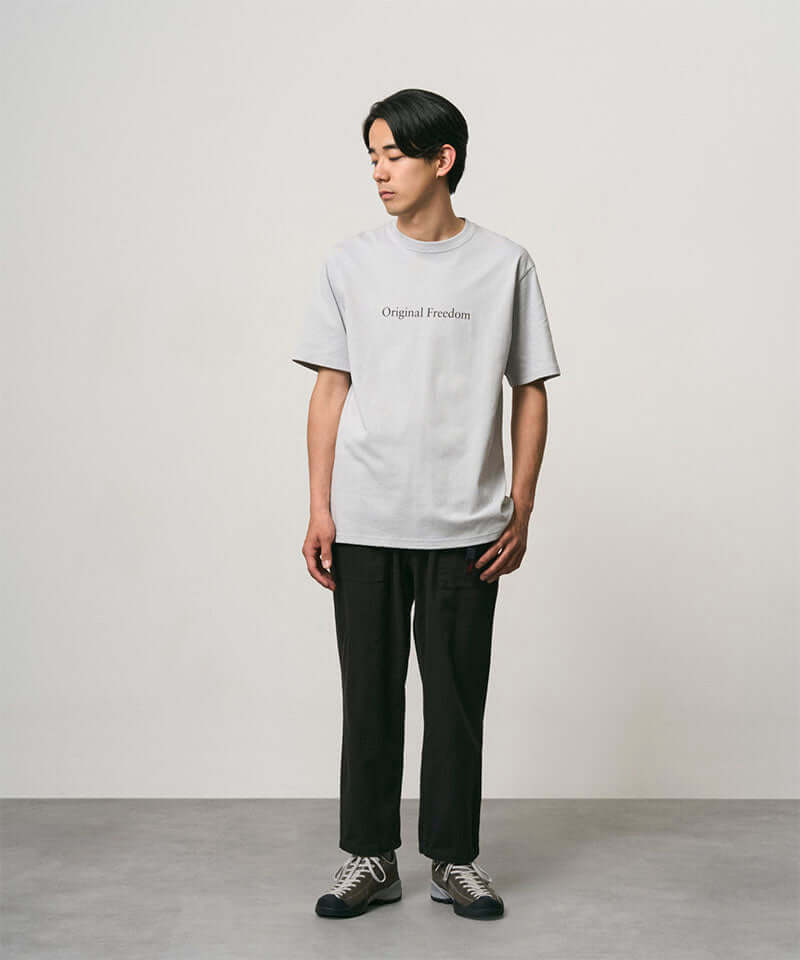Model wearing a mist colored Gramicci Vase Tee and black pants on a light-tone background.