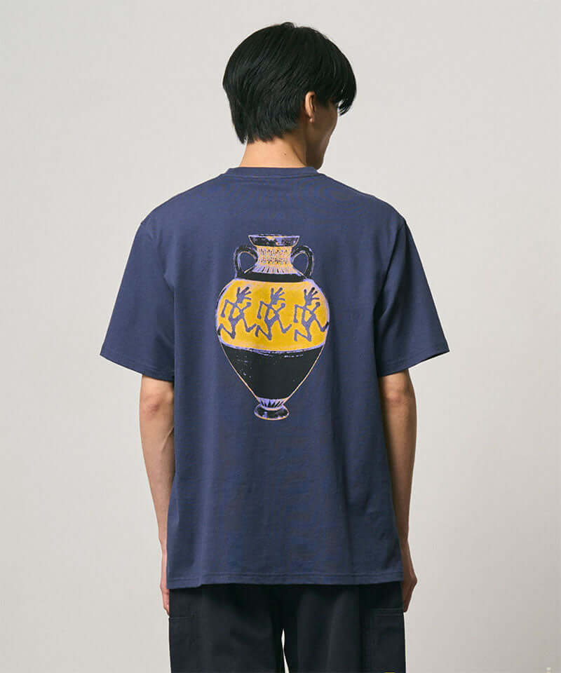 Back view of a model wearing a Gramicci navy Vase Tee with a graphic against a light-tone background.