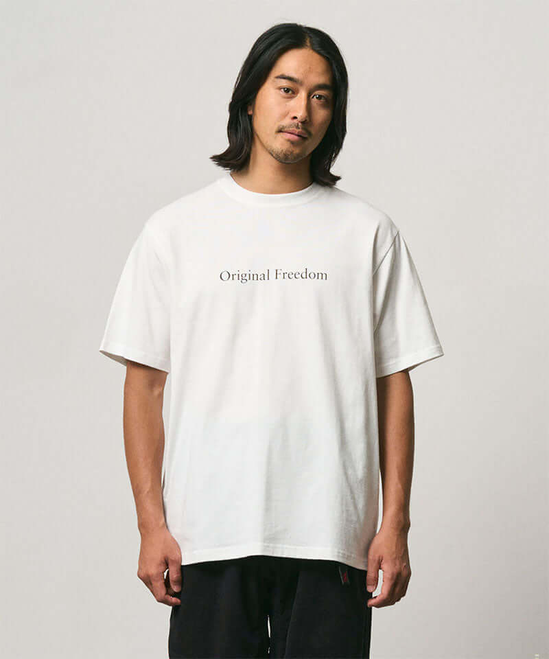 Model wearing a Gramicci white Vase Tee with a graphic against a light-tone background.