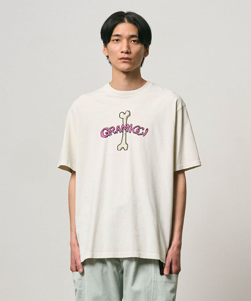 Model wearing a Gramicci natural pigment colored Bone Tee Pigment Dyed with a graphic against a light-tone background.