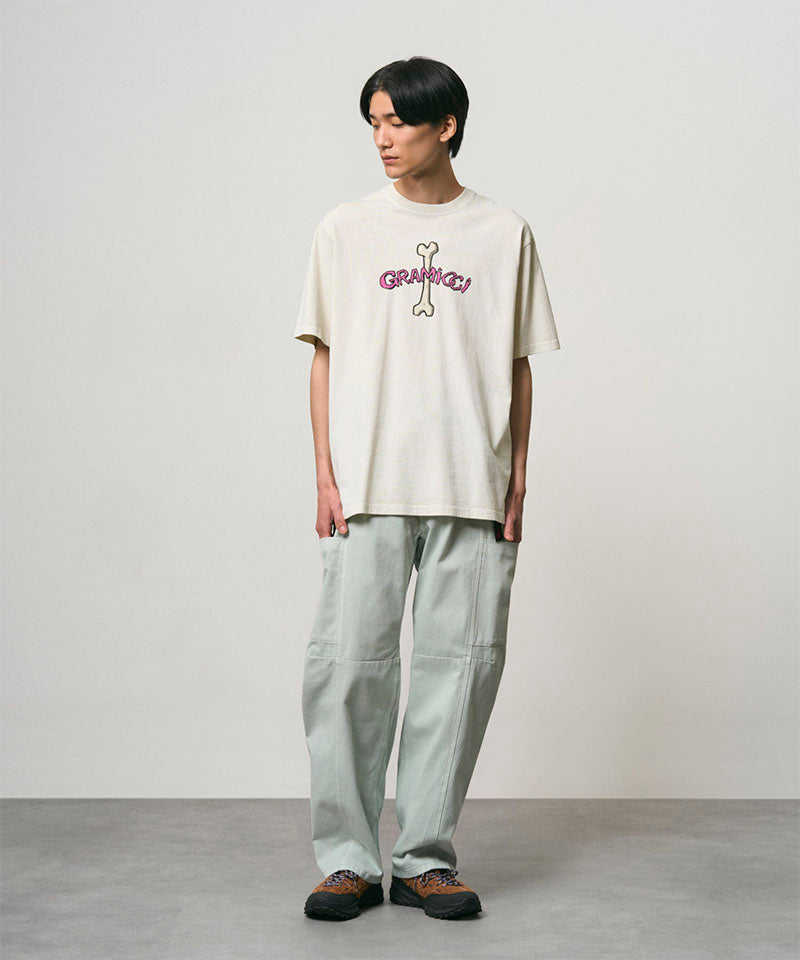 Model wearing a natural pigment colored Gramicci Bone Tee Pigment Dyed with a graphic and light green pants on a light-tone background.