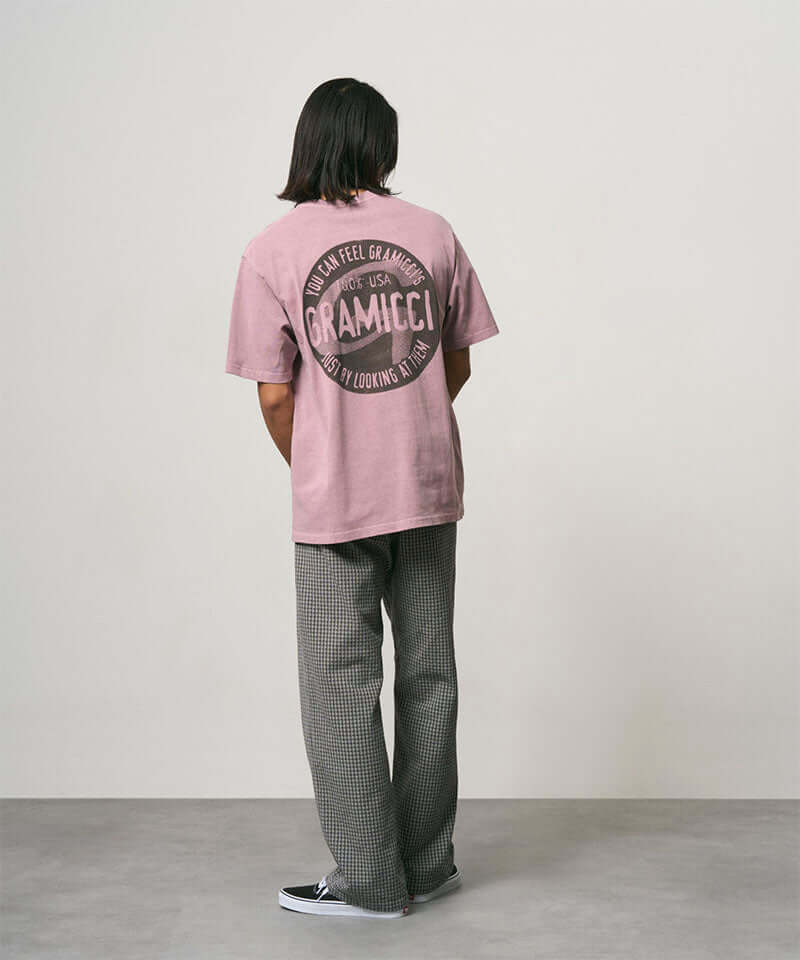 Back view of a model wearing a lilac pigment colored Gramicci Icon Tee Pigment Dyed with a graphic and grey pants on a light-tone background.
