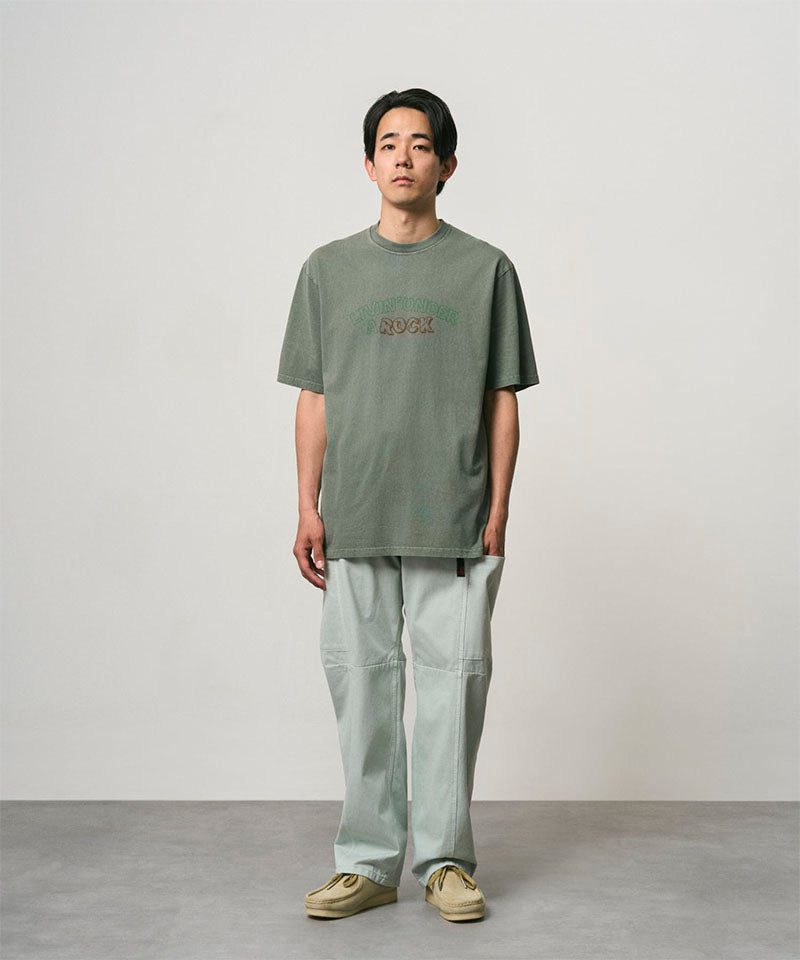 Model wearing a sage pigment colored Gramicci Livin' Tee Pigment Dyed with a Livin' Under A Rock graphic and light green pants on a light-tone background.