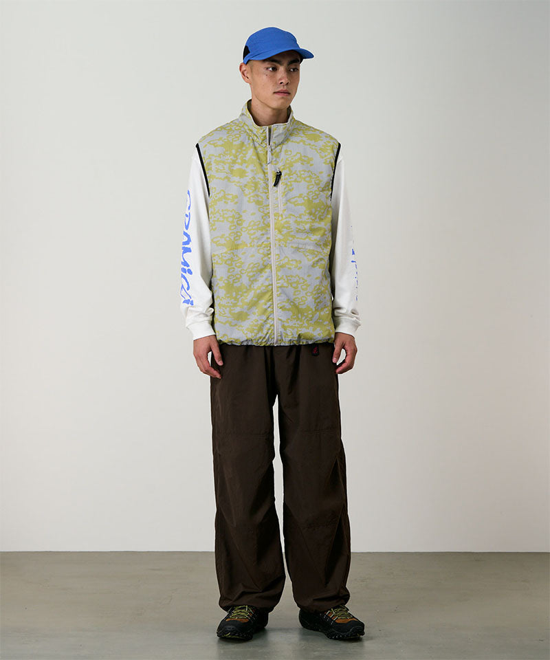 Model wearing a yellow and grey grid camo Gramicci Flyweight Vest and brown pants on a light-tone background.