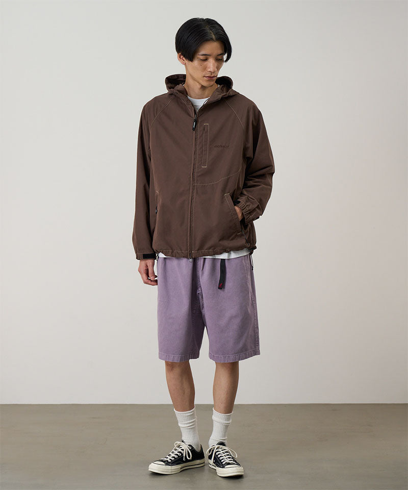 Model wearing a walnut colored Gramicci Milton Hooded Jacket and purple shorts on a light-tone background.