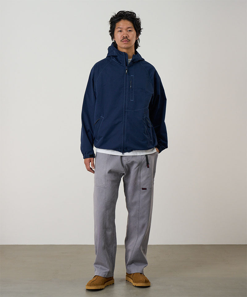 Model wearing a natural dyed grey Gramicci Gadget Pants and blue jacket on a light-tone background.