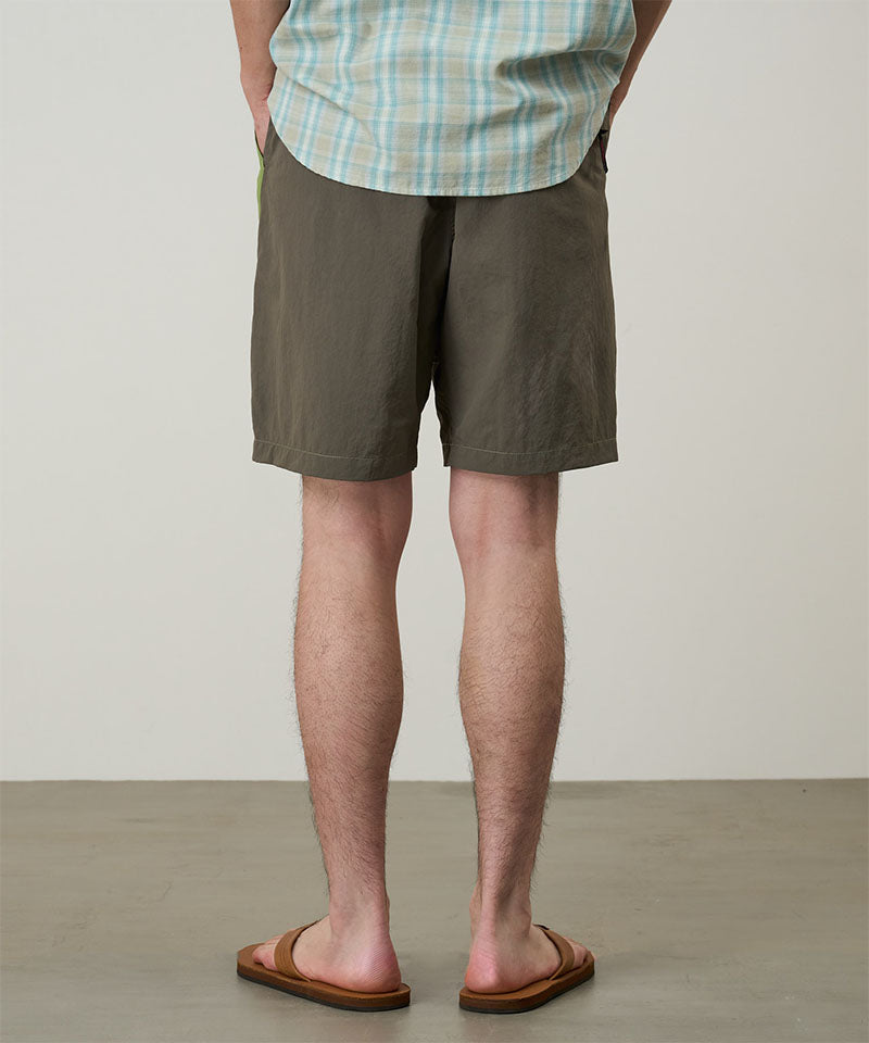 Back view of a model wearing jade green Gramicci Nylon Block Shorts and a green plaid shirt on a light-tone background.