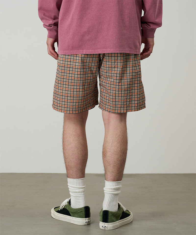 Back view of a model wearing brick check colored Gramicci Checkered Shell Shorts and a red shirt on a light-tone background.