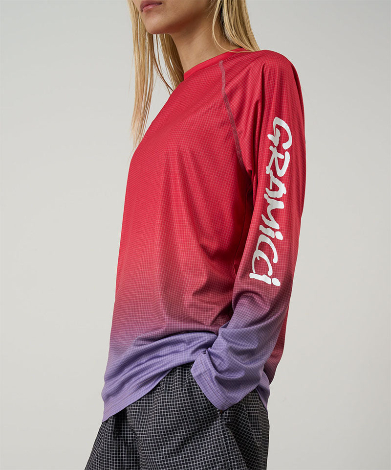 Model wearing a red gradient Gramicci UPF Shield S/S Top with a sleeve graphic and black shorts on a light-tone background.