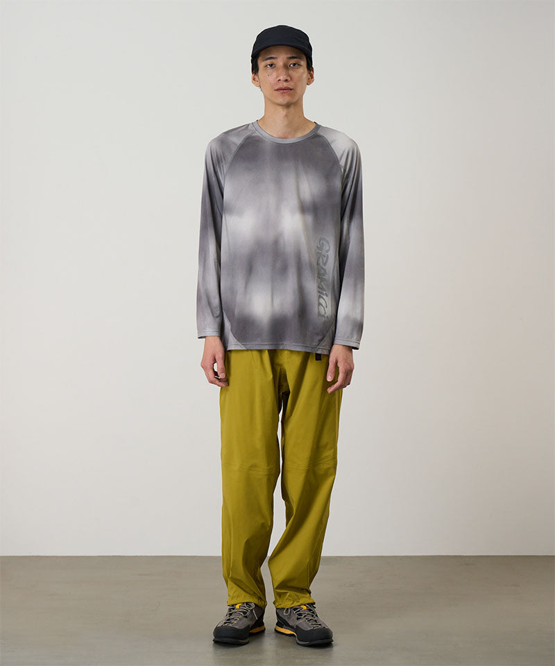 Model wearing a black tie-dye Gramicci L/S 1st Layer shirt with grey graphic and yellow pants on a light-tone background.