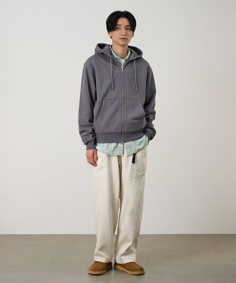 Model wearing a charcoal Gramicci G-Sail Zip Hooded Sweatshirt with a light grey graphic and light colored pants on a light-tone background.