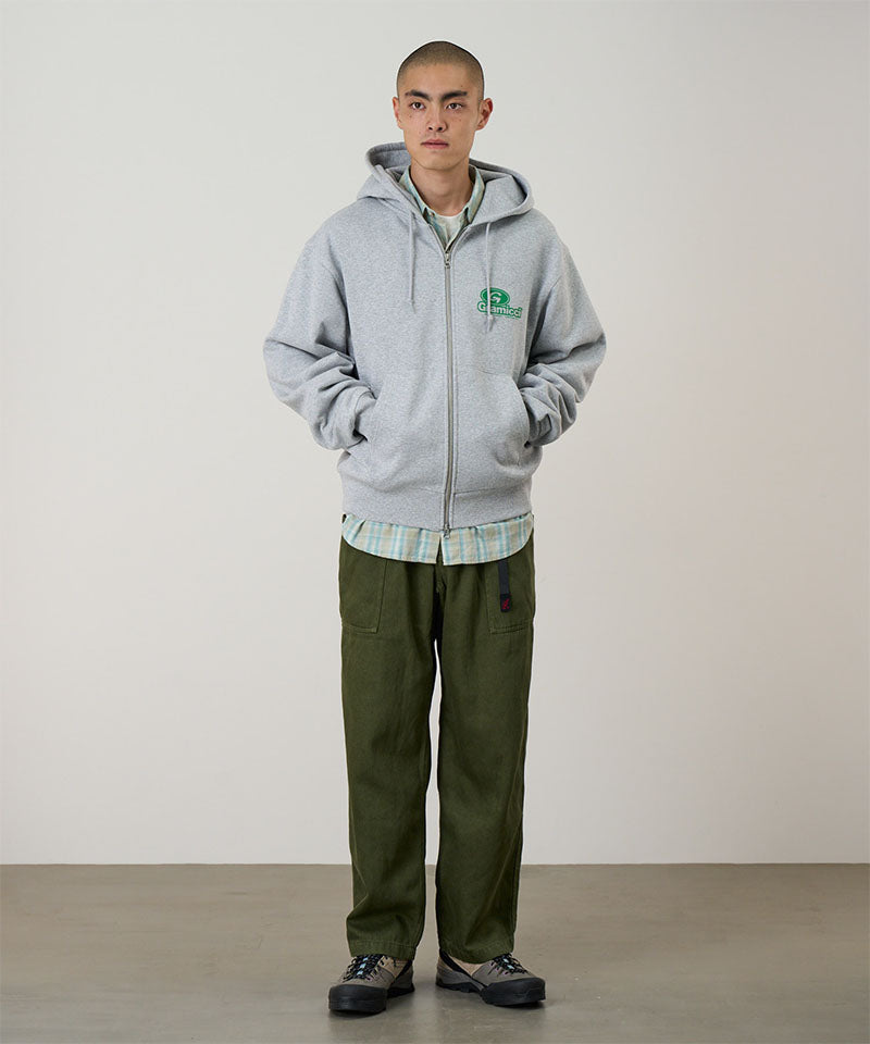 Model wearing a heather grey Gramicci '82 Zip Hooded Sweatshirt with a green graphic and green pants on a light tone background.