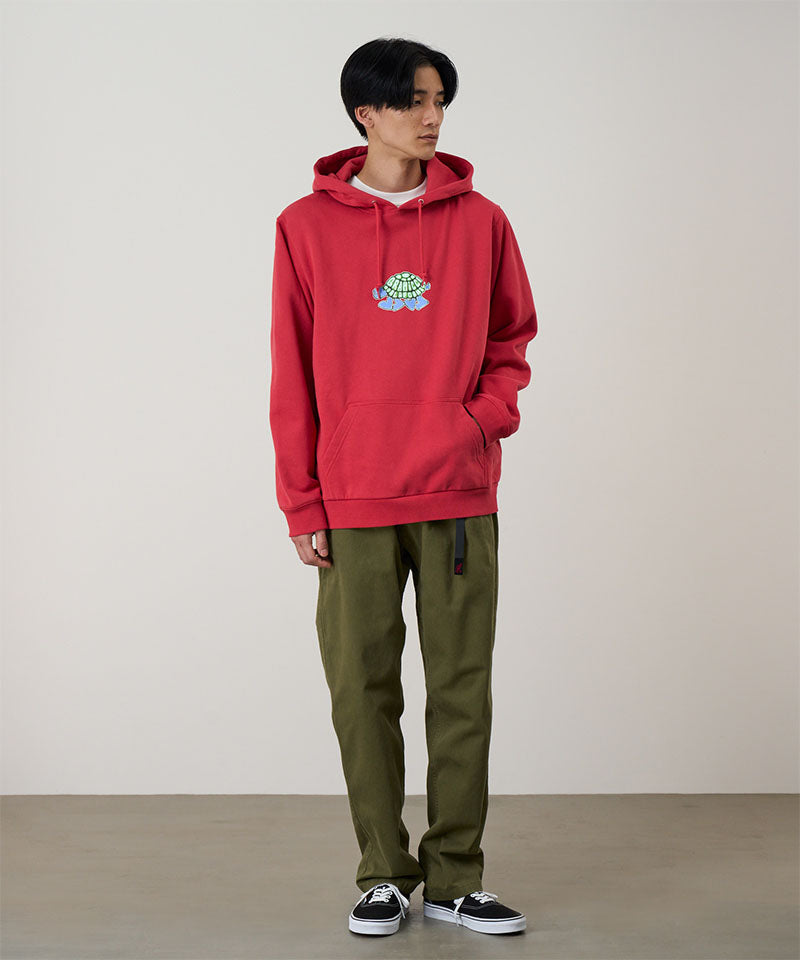 Model wearing a crimson red Gramicci Turtle Hooded Sweatshirt with a turtle graphic and olive pants on a light tone background.