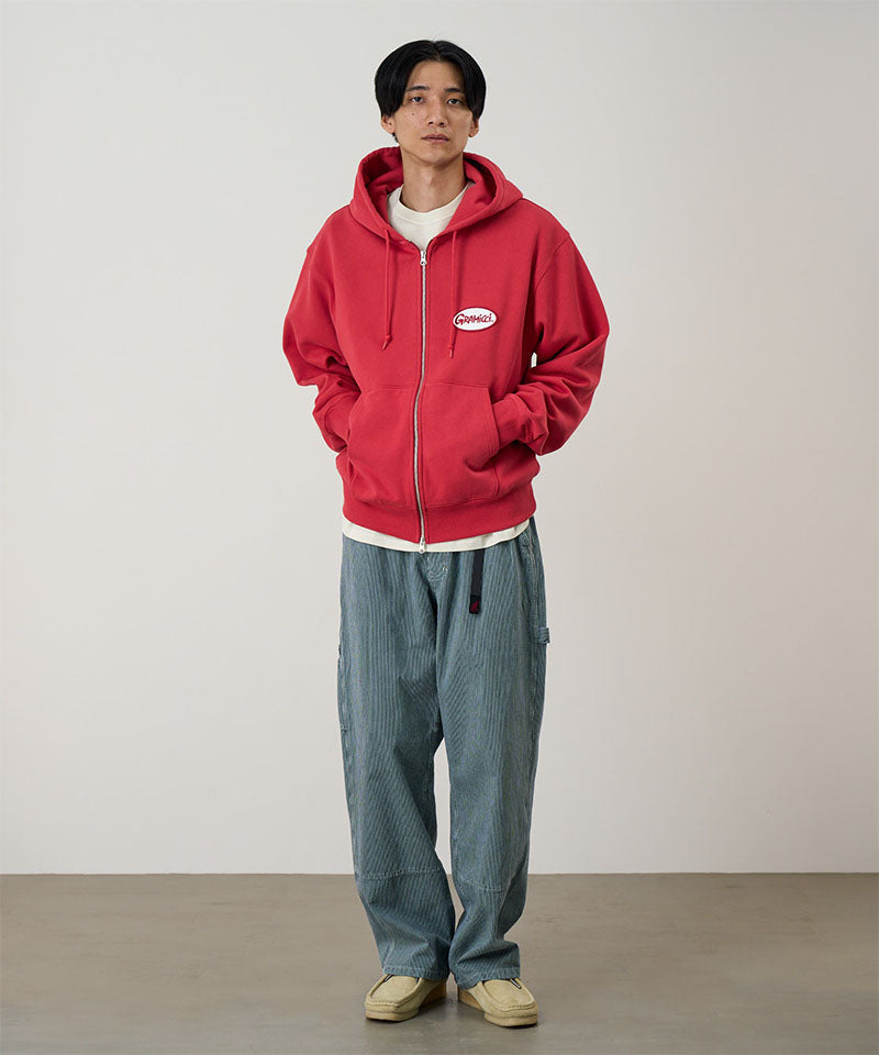Model wearing a crimson red Gramicci Oval Patch Zip Hooded Sweatshirt with a red and white patch and blue jeans on a light-tone background.
