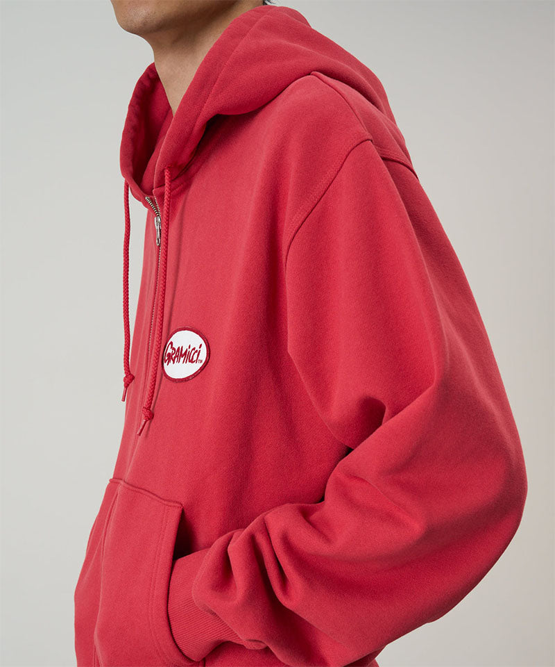 Model wearing a Gramicci crimson red Oval Patch Zip Hooded Sweatshirt with a red and white patch against a light-tone background.