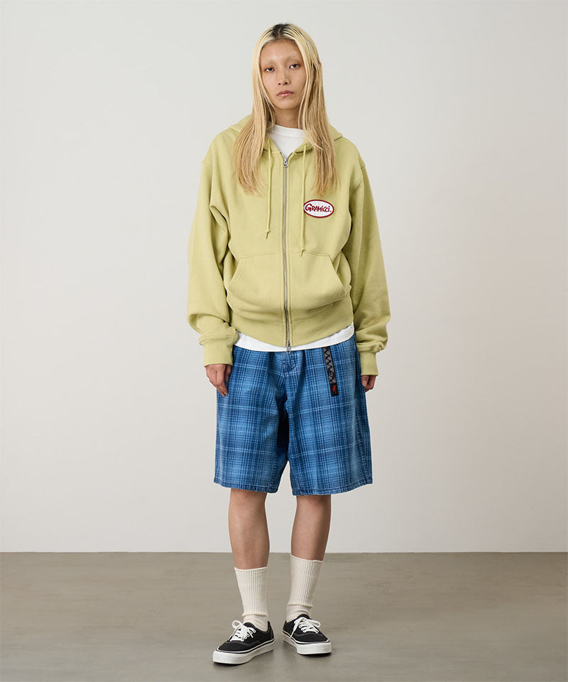 Model wearing a lime Gramicci Oval Patch Zip Hooded Sweatshirt with a red and white patch and blue plaid shorts on a light-tone background.