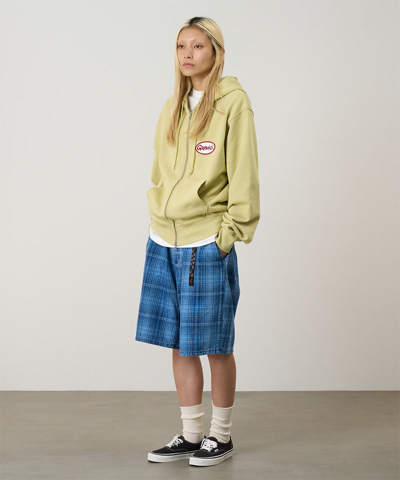 Model wearing a lime Gramicci Oval Patch Zip Hooded Sweatshirt with a red and white patch and blue plaid shorts on a light-tone background.