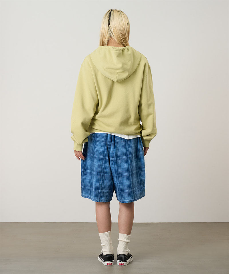 Back view of a model wearing a lime Gramicci Oval Patch Zip Hooded Sweatshirt and blue plaid shorts on a light-tone background.