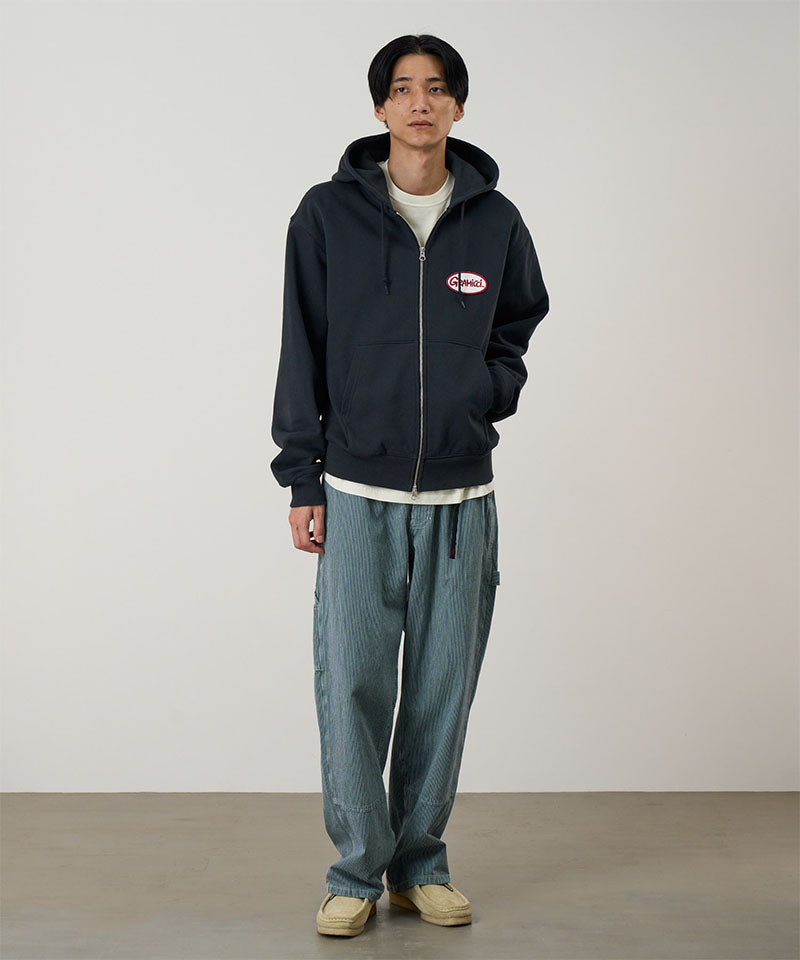 Model wearing a vintage black Gramicci Oval Patch Zip Hooded Sweatshirt with a red and white patch and blue jeans on a light-tone background.
