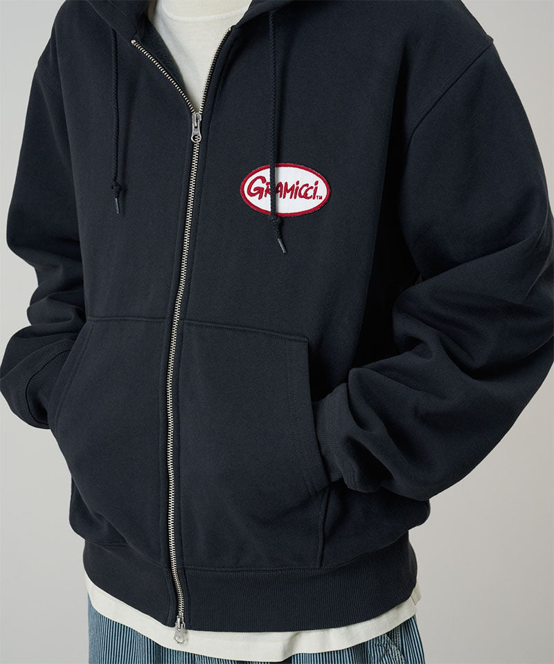 Model wearing a Gramicci vintage black Oval Patch Zip Hooded Sweatshirt with a red and white patch against a light-tone background.