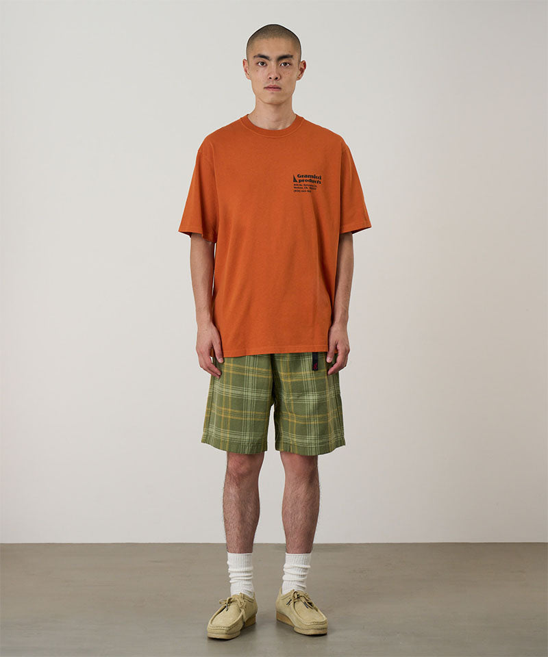 Model wearing a orange Gramicci G-Sail Tee with black graphic and green plaid pants on a light-tone background.