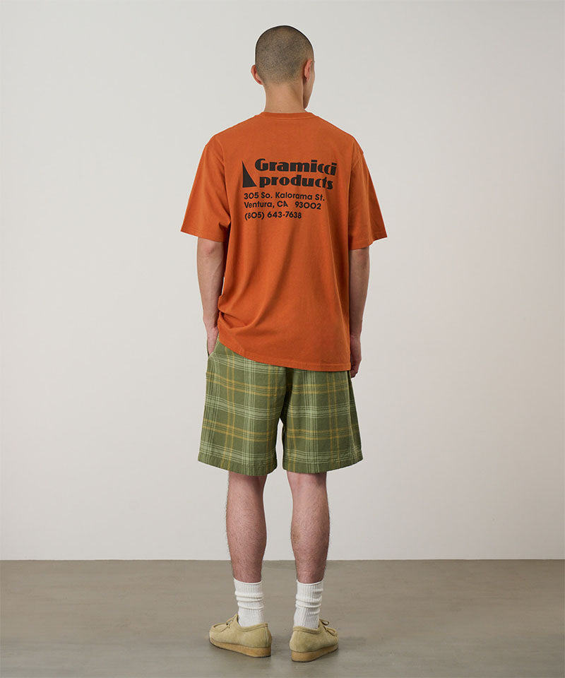Back view of a model wearing a orange Gramicci G-Sail Tee with black graphic and green plaid pants on a light-tone background.