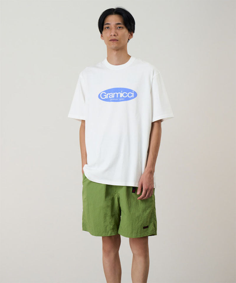 Model wearing a white Gramicci Outdoor Gear Tee with a blue graphic and green shorts on a light-tone background.
