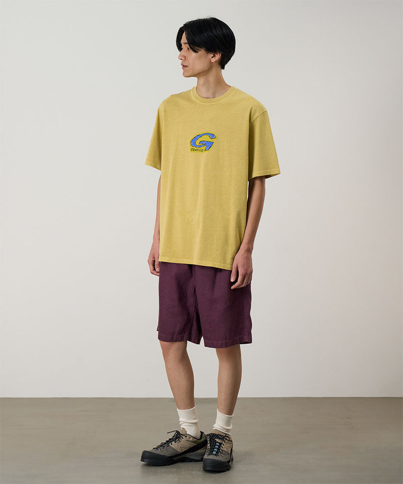 Model wearing a sulpher Gramicci G-Scribble Tee Pigment Dyed shirt with a blue graphic and purple shorts on a light tone background.
