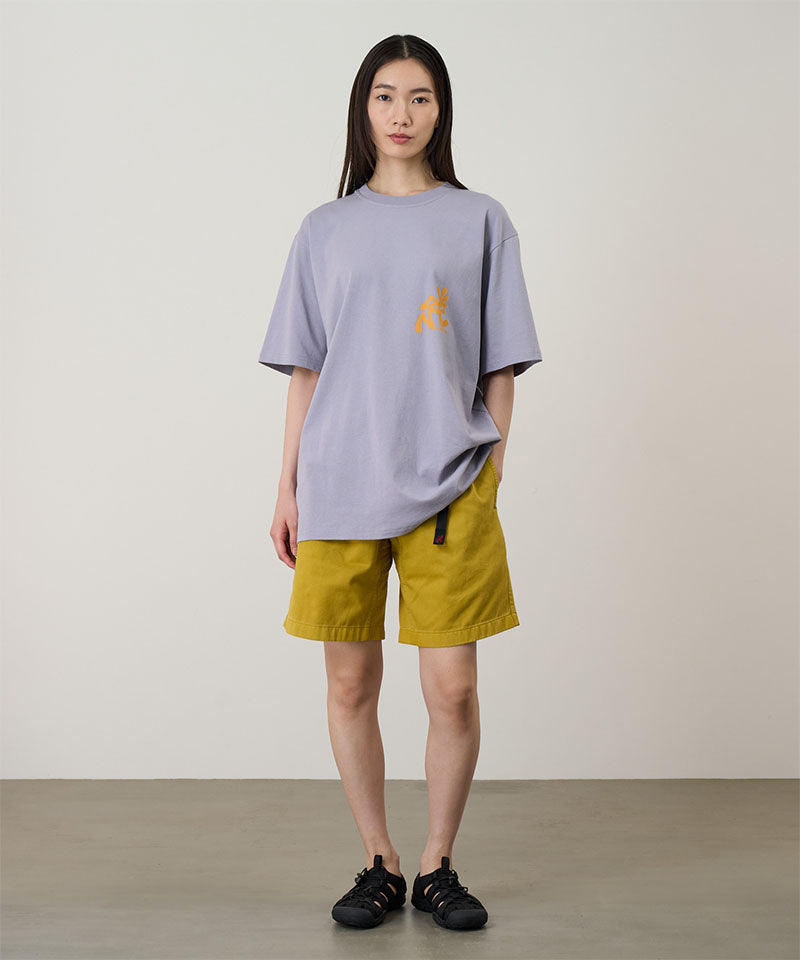 Model wearing a lavender Gramicci Running Man Tee with dark mango colored graphic and yellow shorts on a light-tone background.
