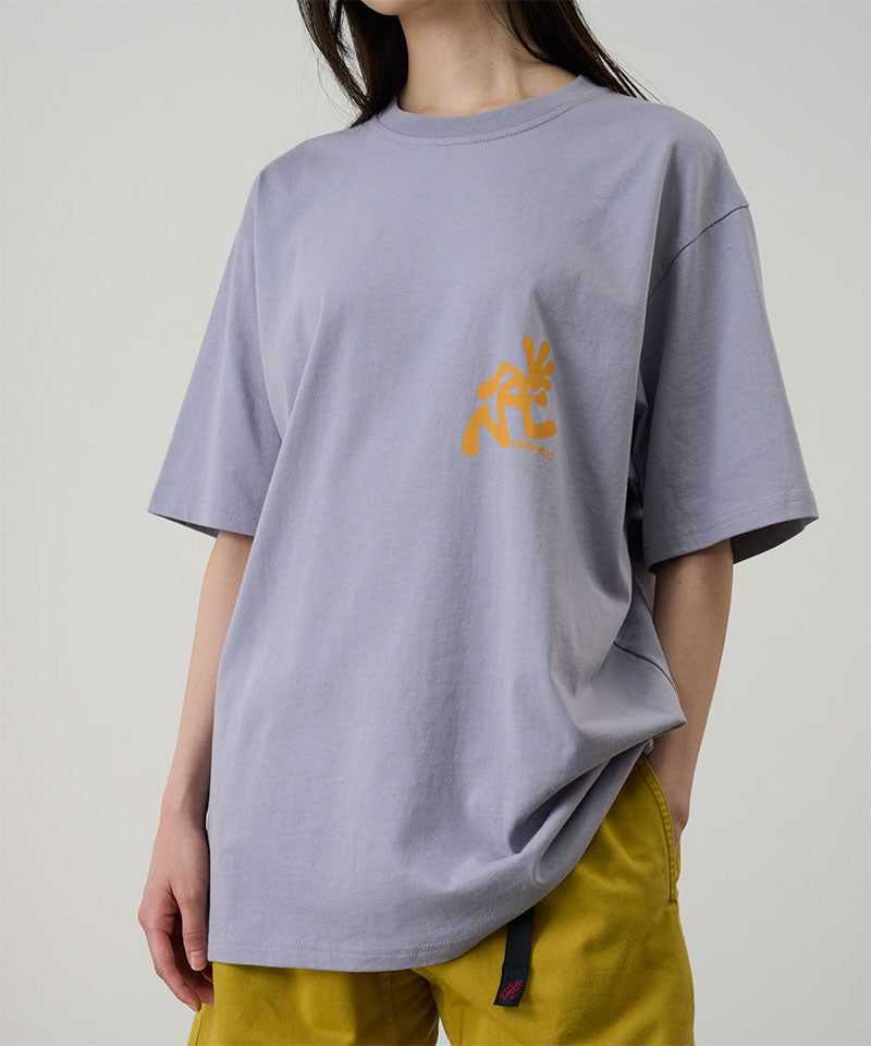 Model wearing a Gramicci lavender Running Man Tee with dark mango colored graphic against a light-tone background.