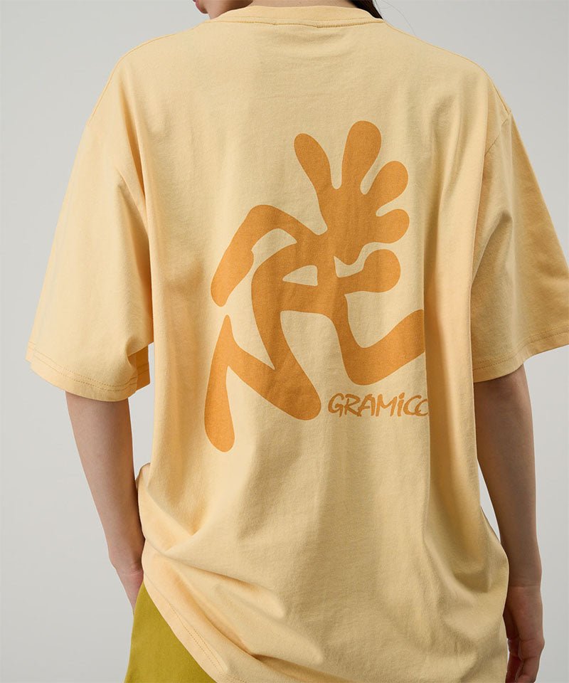 Back view of a model wearing a Gramicci mango colored Running Man Tee with dark mango colored graphic against a light-tone background.