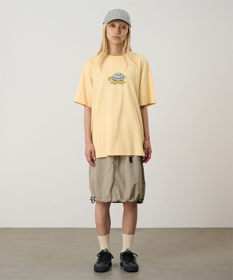 Model wearing a mango Gramicci Turtle Tee with a a turtle graphic and beige skirt on a light tone background.