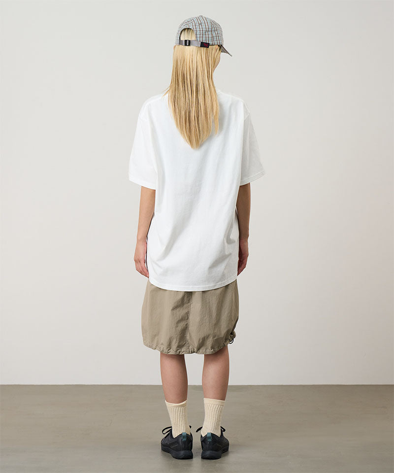 Rear view of a model wearing a white Gramicci Turtle Tee and beige skirt on a light tone background.