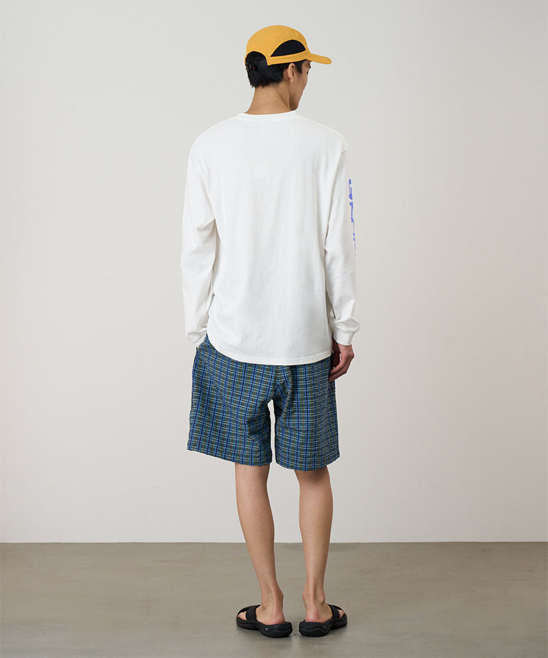 Back view of a model wearing a white Gramicci Freedom L/S Tee with a blue graphic on sleeves and blue checkered shorts on a light tone background.