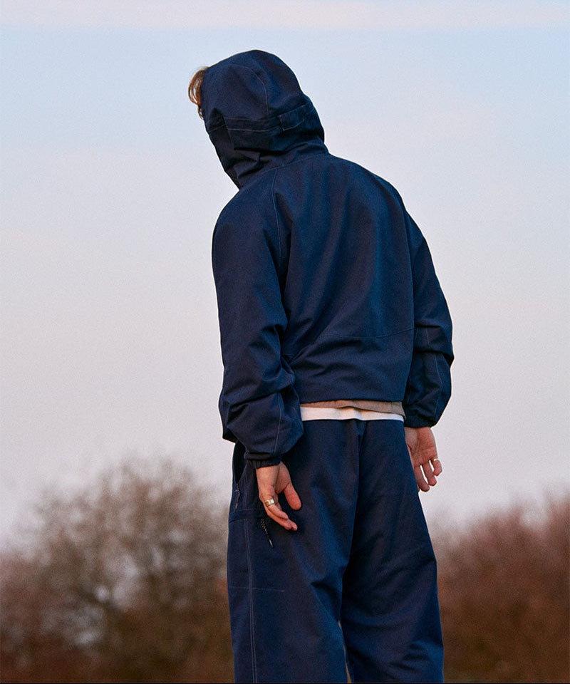 Man wearing a navy blue Gramicci rain jacket and pants standing outdoors with a blurred background