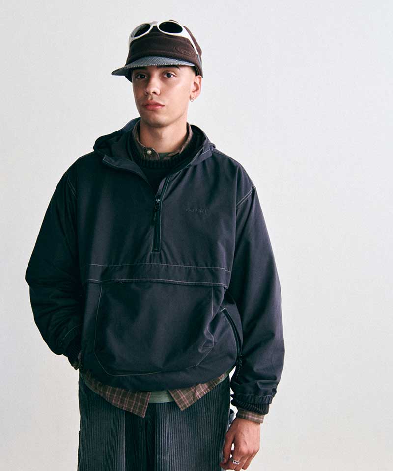 Model wearing a Gramicci black hooded half-zip anorak layered over a plaid shirt, dark corduroy pants, and a fleece brimmed cap with sunglasses.