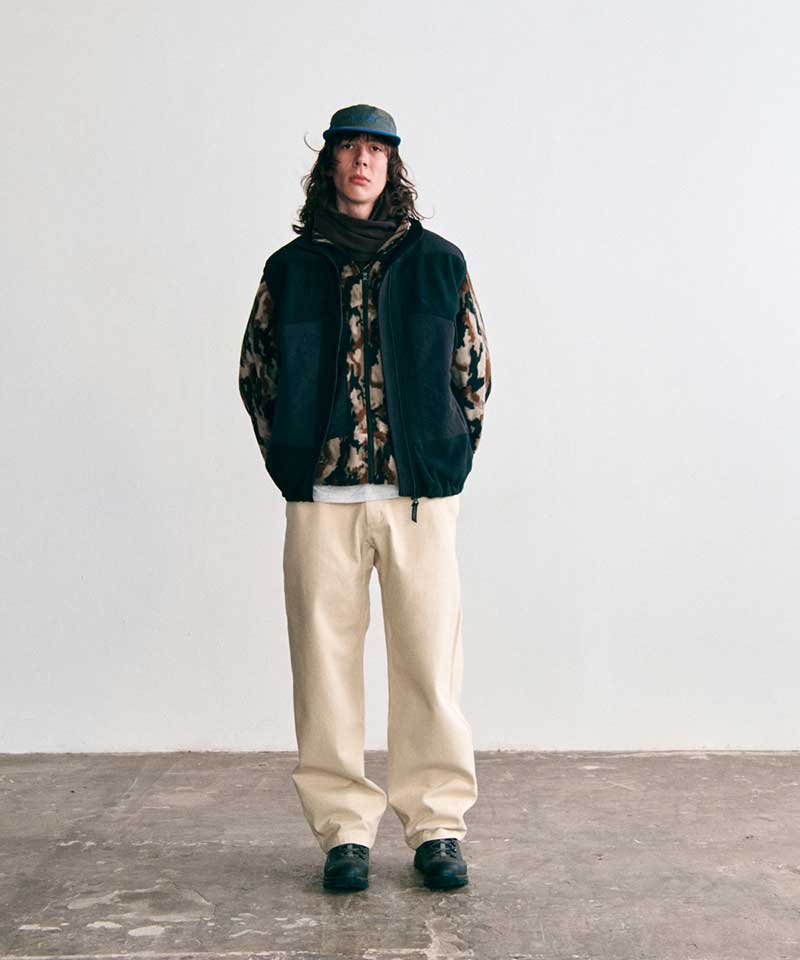 Model wearing a Gramicci black fleece vest layered over a camo fleece jacket, cream relaxed pants, a dark neck gaiter, and a cap.