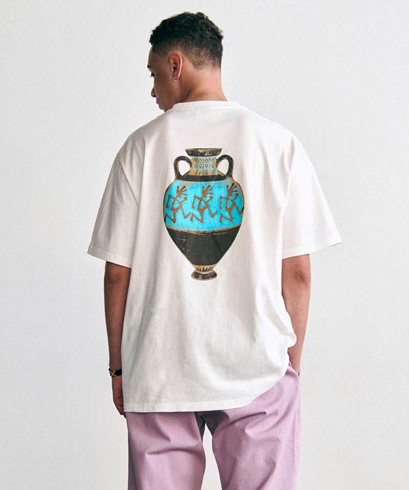 Gramicci white T-shirt with a turquoise vase graphic featuring the Running Man logo on the back, paired with lilac pants and worn by a person standing against a white background.
